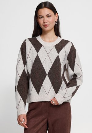 Femme portant un pull beige et marron à motif losanges, manches longues, et un pantalon marron sur un fond clair uni.
