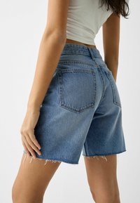 Shorts in denim blu con orlo sfilacciato, design a cinque tasche e vita media, che mostrano una texture liscia e uno stile classico.