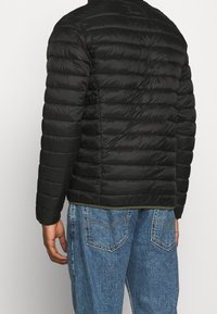 Giacca a vento nera con design trapuntato, maniche lunghe e colletto alto. Realizzata in materiale leggero. Indossata sopra jeans con texture.