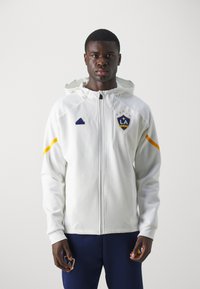 Witte zip-up jacket met capuchon, voorzien van een blauw Adidas-logo en LA Galaxy-embleem, geaccentueerd met gele strepen op de mouwen.