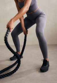 Gray athletic leggings and matching top, textured fabric, holding thick black battle ropes, black sneakers on a light gray floor.