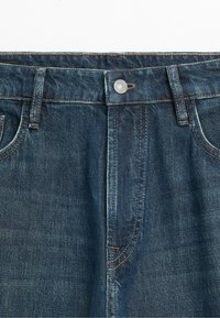 Detail modrých džínů vpředu s kovovým knoflíkem, poutky na pásek a viditelným prošitím na kapsách a švech.