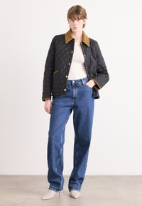 Jachetă neagră matlasată cu guler maro, buzunare frontale și accesorii aurii, asociată cu blugi din denim cu talie înaltă de culoare albastră și pantofi bej cu vârf ascuțit.