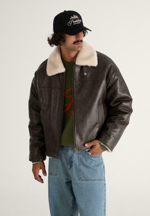 Braune Lederjacke mit einem weißen Schafsfellkragen, Reißverschluss und Seitentaschen; getragen über einem grünen Pullover und kombiniert mit hellblauen Jeans.