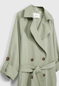 Gabardina verde claro hecha de tela suave, con un diseño de doble botonadura, gran cuello, botones marrones y un cinturón atado a la cintura.