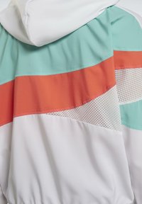 Giacca a vento colorata con base bianca, con accenti in verde menta, arancio corallo e rete. Il design include un blocco di colori geometrici.