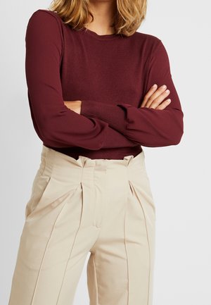 Femme portant un haut bordeaux à manches longues avec des poignets côtelés et un pantalon beige plissé taille haute, les bras croisés.