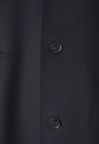 Dettaglio del cappotto in misto lana blu navy, caratterizzato da una texture liscia, due bottoni frontali e un taglio su misura con cuciture precise.