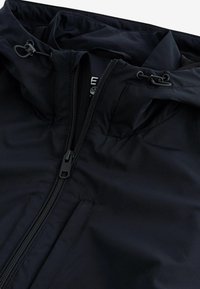 Veste noire avec une fermeture éclair frontale, des cordons réglables sur la capuche et une poche poitrine avec une fermeture éclair dissimulée.