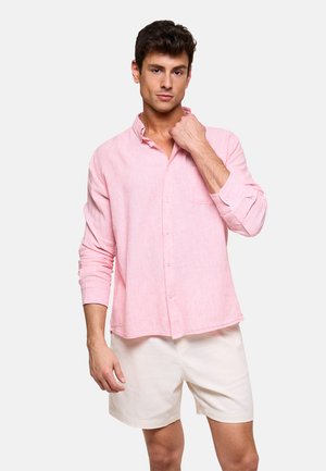 Joven con camisa de manga larga de color rosa claro y pantalones cortos beige, posando con una mano cerca del cuello.