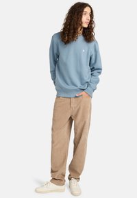Sudadera de color azul claro en material suave con cuello redondo y logotipo. Combinada con pantalones de pana beige y zapatillas blancas. Ajuste casual.