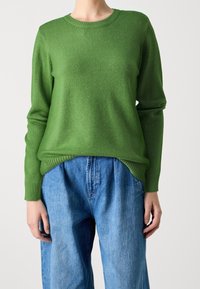 Grüner Strickpullover mit rundem Halsausschnitt, langen Ärmeln und gerippten Manschetten, kombiniert mit hellblauen, locker sitzenden Jeans.