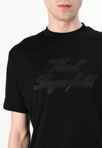Juodas medvilninis marškinėlis su trumpomis rankovėmis, kuriame centre išsiuvinėtas pakeltas juodas tekstas "Karl Lagerfeld". Sklandžią tekstūrą turintis, apvalaus kaklo dizainas.