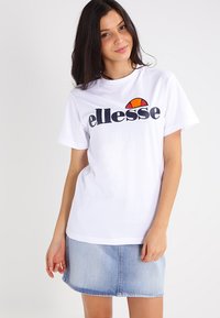 Hvit bomull t-skjorte med marineblå "ellesse"-logo, med en fargerik halvmåneaksent. Modellen har på seg et lyst denimskjørt.