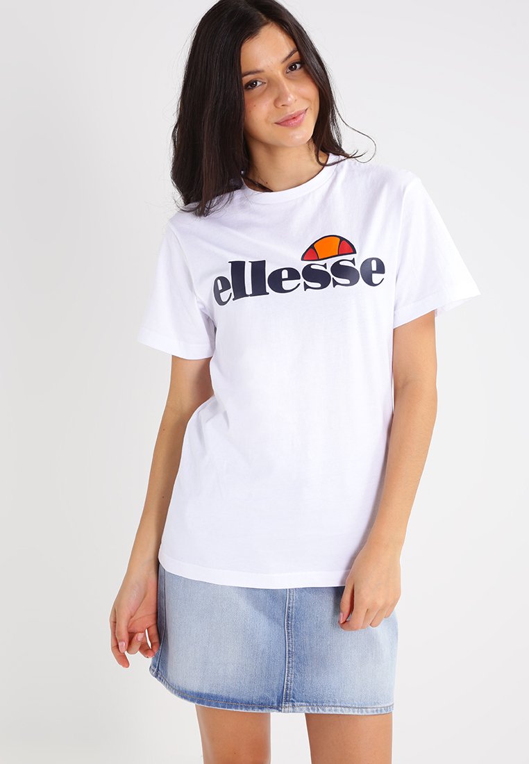 Hvit bomull t-skjorte med marineblå "ellesse"-logo, med en fargerik halvmåneaksent. Modellen har på seg et lyst denimskjørt.
