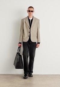 Blazer beige, camicia nera e jeans scuri. Borsa nera e scarpe nere. Modello indossa occhiali da sole, in piedi contro uno sfondo bianco uniforme.