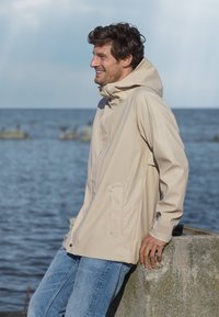 Beige vattentät jacka med hög krage, dragkedja framtill och sidofickor. Materialet verkar slätt. Bärs över blå jeans.