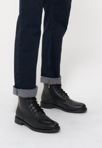 Czarne skórzane botki za kostkę o gładkim wykończeniu, zawiązywane, z bocznym zamkiem. Połączone z podwiniętymi ciemnoniebieskimi dżinsami.