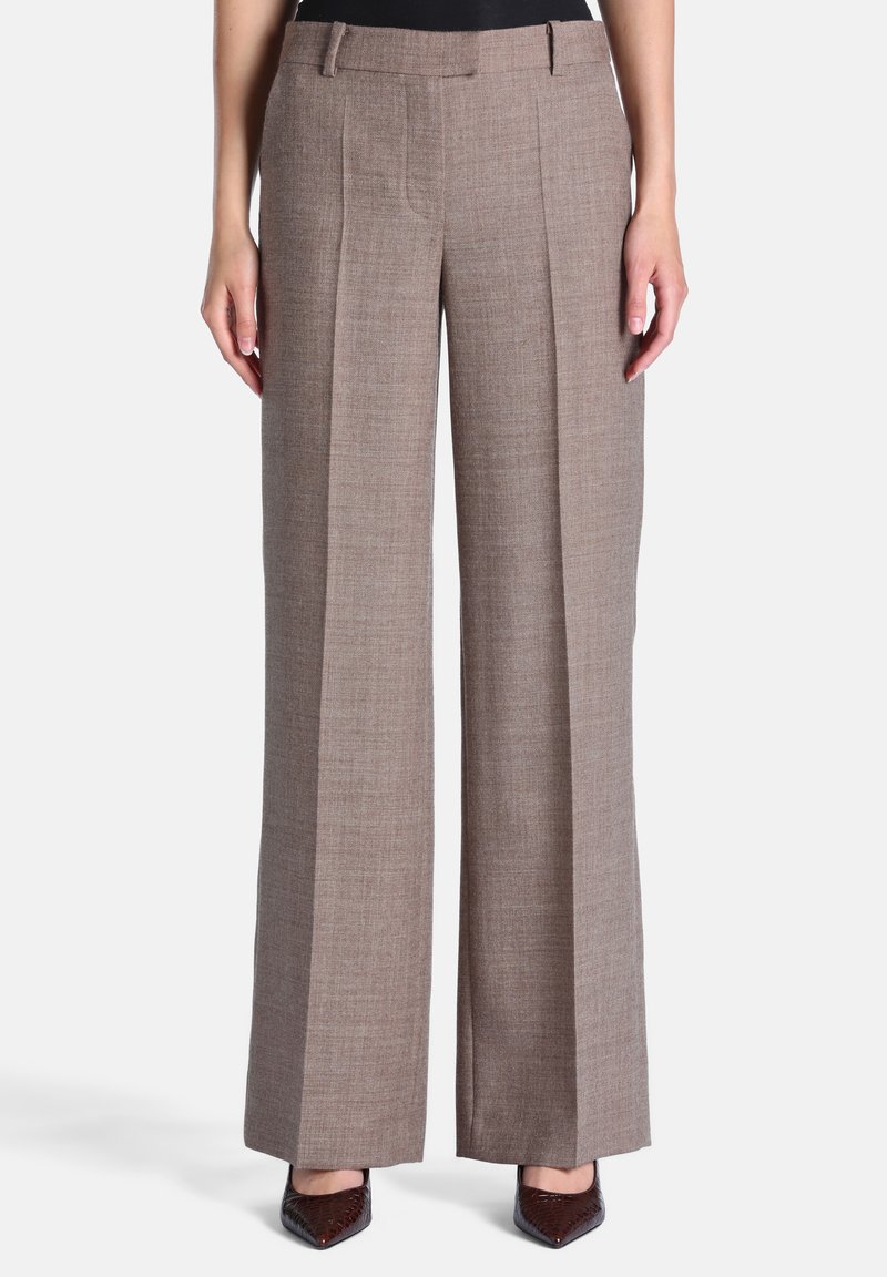 Femme portant un pantalon taupe à jambes larges avec plis devant et des talons marron à bout pointu, debout devant un fond clair uni.