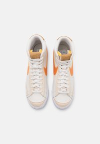 Höga sneakers med en ovandel i krämfärgad och ljusbrun läder, vita snören och en orange swoosh på sidan, med en texturerad tunga och krage.