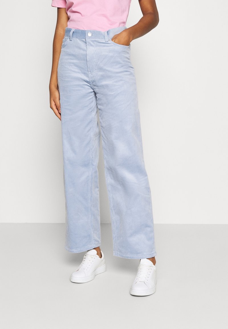Personne portant un pantalon large en velours côtelé bleu clair, une chemise rose et des baskets blanches, une main dans la poche.