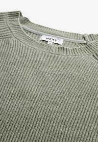 Olijfgroene ribgebreide trui met een ronde hals. Bevat een label met de tekst "NEXT" en maat "M" aan de binnenkant. De zachte textuur is zichtbaar in de stof.