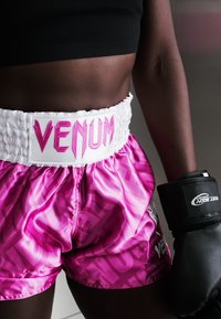 Πυγμάχος φοράει ροζ σορτς Venum και μαύρο αθλητικό τοπ, με μαύρο γάντι πυγμαχίας στο δεξί χέρι, στέκεται σε γυμναστήριο.
