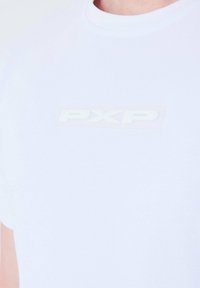 Camiseta blanca de algodón con un logo "PXP" texturizado y transparente en el pecho. Cuello redondo y mangas cortas. Tela suave.