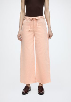 Γυναίκα που φοράει φαρδιά παντελόνια σε ροδακινί απόχρωση με κορδόνι στη μέση, σκούρο καφέ αμάνικο τοπ και σκούρα καφέ δερμάτινα μοκασίνια.