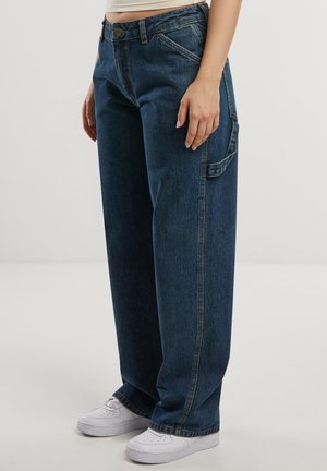 Person iført løse mørkeblå denimjeans med sidelomme detalje og hvide sneakers, stående mod en ensfarvet lys baggrund.