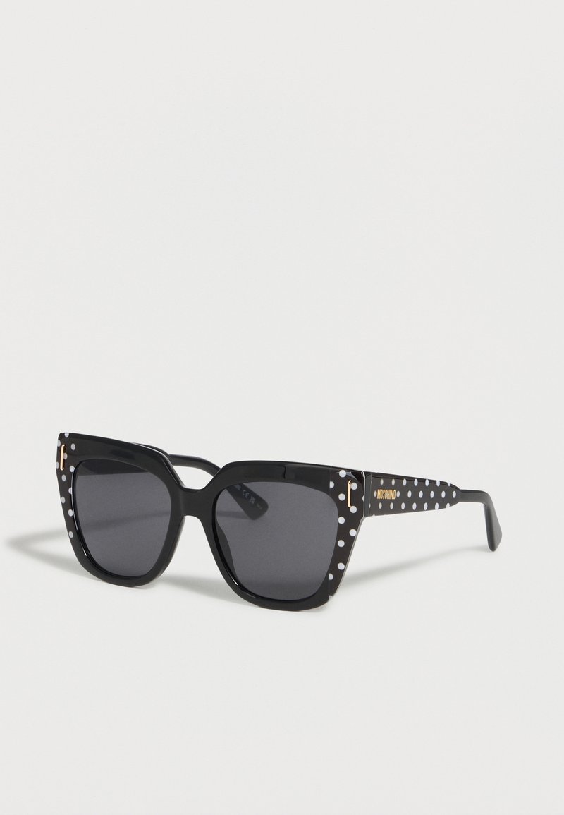 Lunettes de soleil carrées noires avec des verres foncés et des pois blancs sur la monture et les branches, avec un petit logo Moschino doré sur le côté.