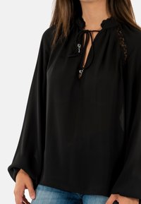 Femme portant un haut noir ample avec des détails en dentelle et un col à nouer, associé à un jean bleu, vue des épaules aux hanches.
