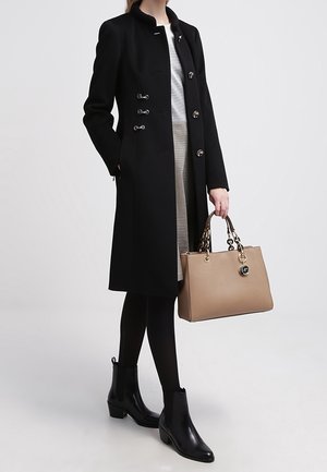 Frau trägt einen schwarzen knielangen Mantel, schwarze Stiefeletten und hält eine beige Handtasche mit goldenen Kettenhenkeln.