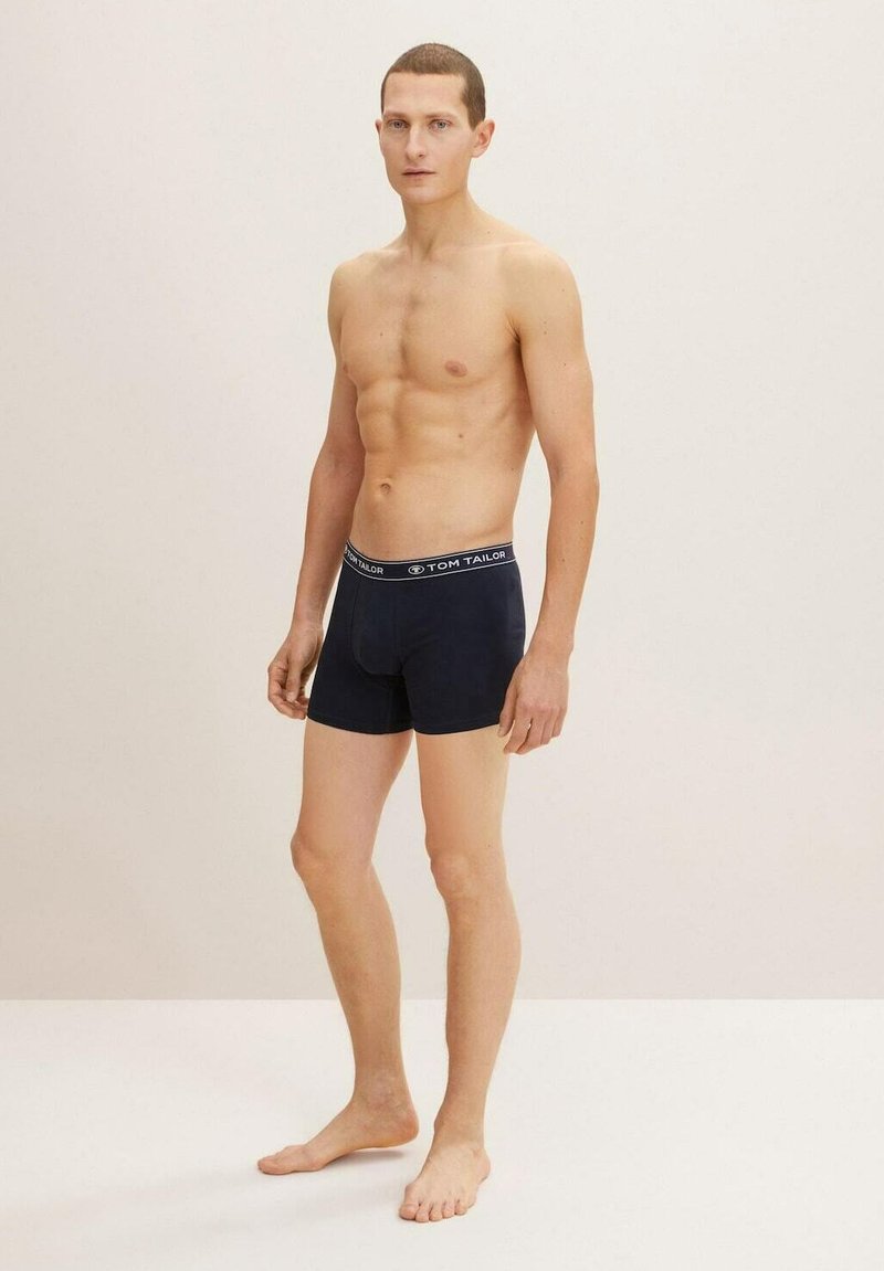 Dunkelblaue Boxershorts mit einem schwarzen Bund, der mit weißem "TOM TAILOR"-Branding versehen ist, aus einem glatten, dehnbaren Stoff. Das Modell steht barfuß.