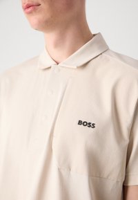 Šviesiai ruda polo marškinėliai su struktūrizuotu apykakle, turintys tekstūruotą panelę ir diskretų juodą "BOSS" logotipą kairėje krūtinės pusėje. Sklandus audinio paviršius.