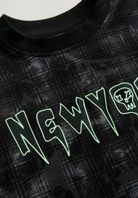 Fekete grafikus kapucnis pulóver, amely texturált kockás mintával rendelkezik. Élénk zöld betűkkel van ellátva, amelyeken a "NEWYORK" felirat és egy koponya dizájn szerepel.