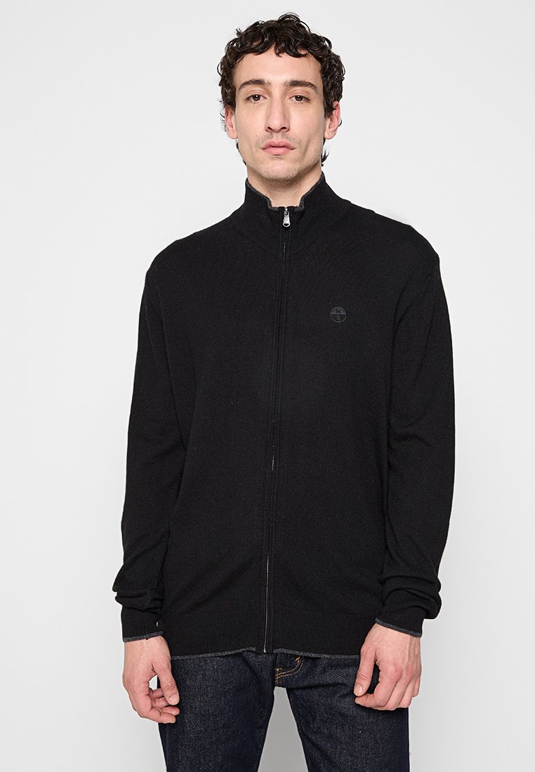 Cardigan negro con cremallera y cuello alto, hecho de un material de punto suave. Presenta un pequeño logo en el pecho y puños y dobladillo acanalados.