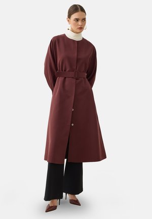Donna che indossa un lungo cappotto bordeaux con cintura sopra un dolcevita bianco, pantaloni neri a gamba larga e décolleté bordeaux con punta, in piedi con le mani dietro la schiena.