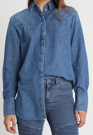 Femme portant une chemise en jean à manches longues rentrée dans un pantalon en jean taille haute avec des détails côtelés sur les cuisses.