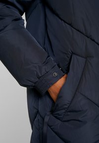 Marine pufferjack met een gewatteerd ontwerp, elastische manchetten en zijzakken. Beschikt over een gladde, glanzende textuur en een trekkoord onderaan.