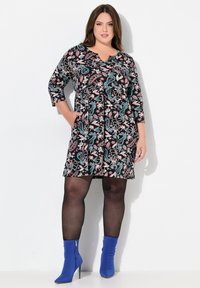 Robe noire avec un motif floral en rose et teal, manches trois quarts, décolleté en V et poches latérales. Portée avec des bottines bleues.