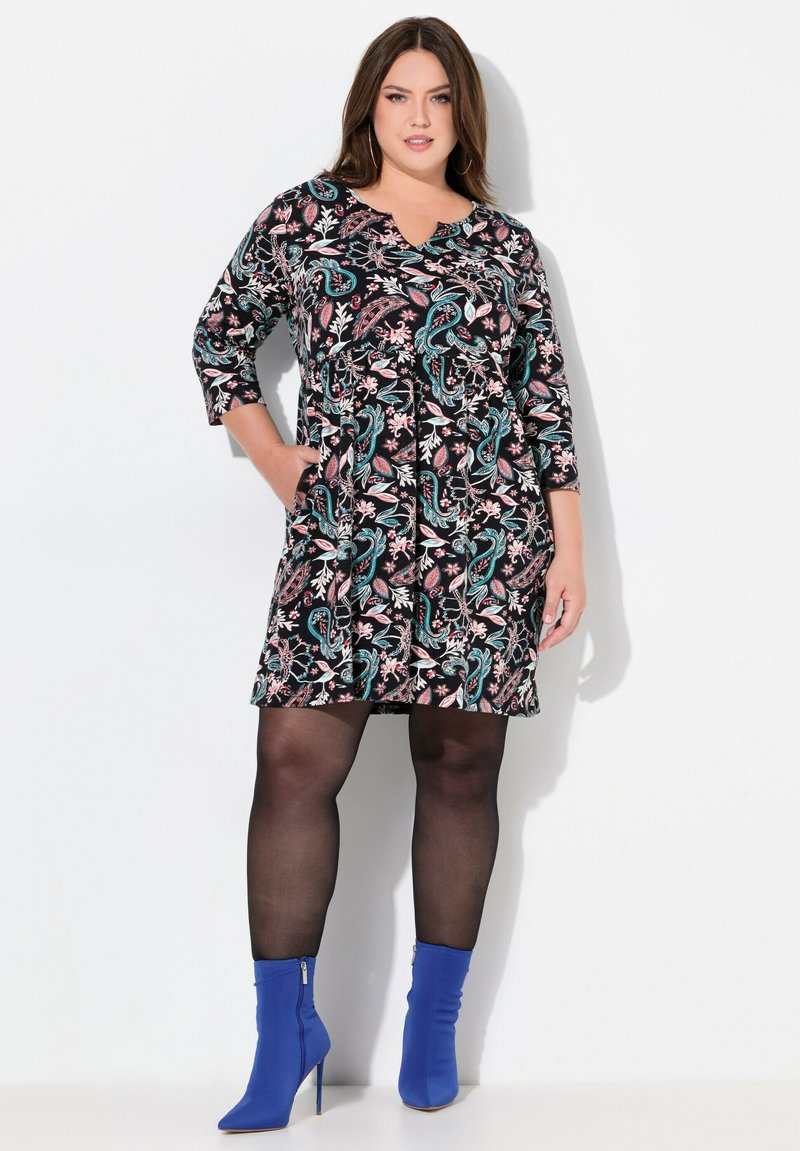 Robe noire avec un motif floral en rose et teal, manches trois quarts, décolleté en V et poches latérales. Portée avec des bottines bleues.