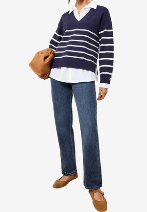 Personne portant un pull à col en V rayé bleu marine et blanc sur une chemise blanche, un jean bleu, des ballerines beige et tenant une pochette beige.
