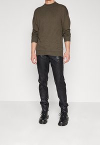 Olivgrön stickad tröja med hög hals, i kombination med åtsittande svarta läderbyxor och svarta snörboots. Slät textur och åtsittande design.