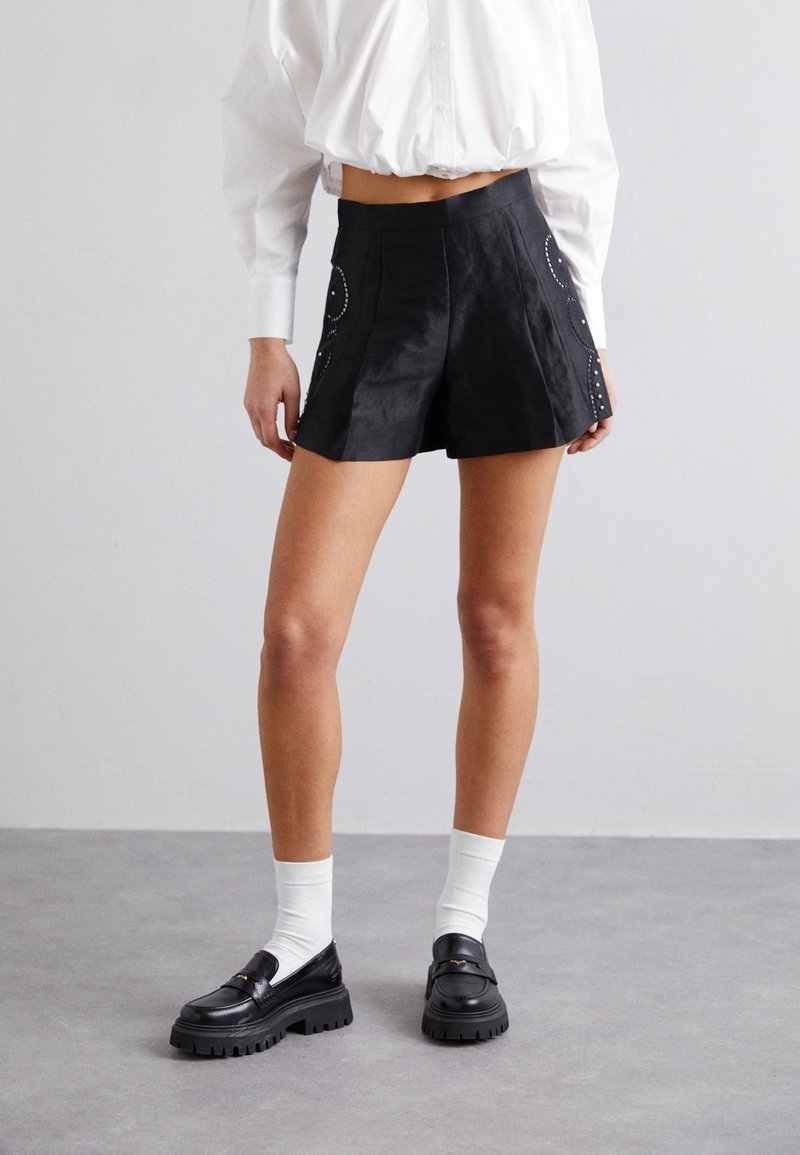 Personne portant un short noir taille haute avec des détails sur les côtés, une chemise courte blanche, des chaussettes blanches et des mocassins noirs épais, debout sur un sol gris.