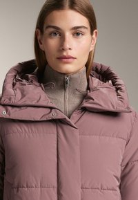 Różowa pikowana kurtka z kapturem, zapinana na zamek z przodu, z elastycznymi wstawkami. Nosi się ją na jasno-brązowym swetrze z wysokim kołnierzem.