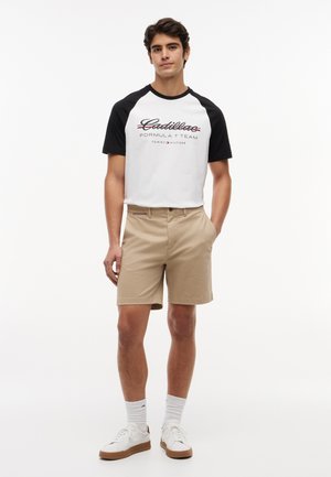 Giovane in piedi, che indossa una T-shirt a maniche corte bianca e nera del team Formula 1 Cadillac, pantaloncini beige, calzini bianchi e scarpe da ginnastica bianche.