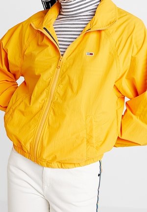 Personne portant une veste zippée jaune vif sur un col roulé rayé noir et blanc, associée à un pantalon blanc avec des bandes latérales.