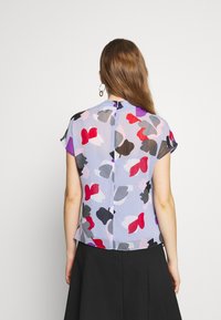 Blusa transparente con un patrón abstracto multicolor en rojo, rosa, gris y púrpura. Cuenta con mangas cortas y un cierre de llave en la parte de atrás.