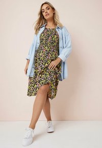 Schwarzes Blumenkleid mit gelben und pinken Blumen, knielang mit hohem Schlitz, kombiniert mit einer hellblauen Jeansjacke und weißen Sneakers.